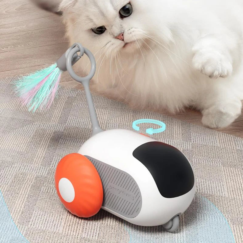 Cat toy resembling a small vehicle with a feather on wheels, next to a cat on a wooden floor.