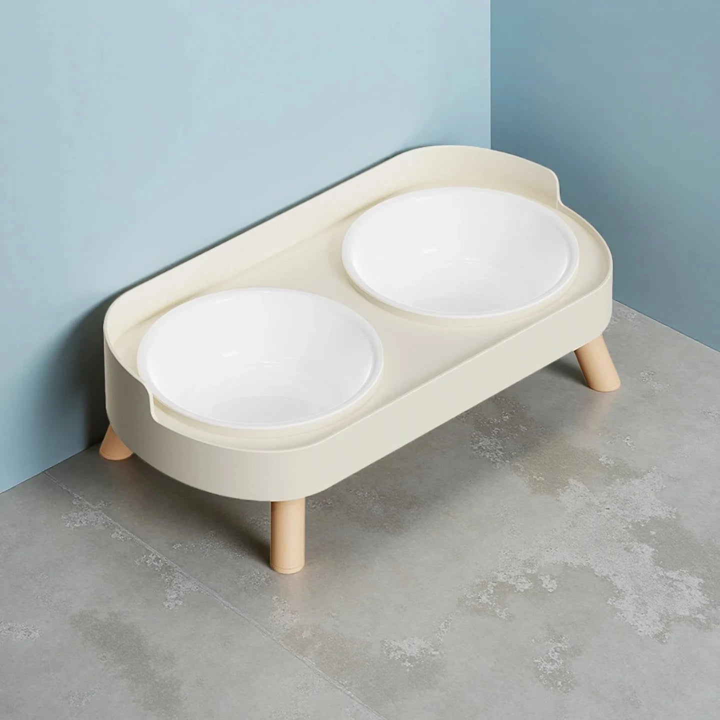 Beige elevated pet feeder with two white bowls on a concrete floor against a light blue wall.